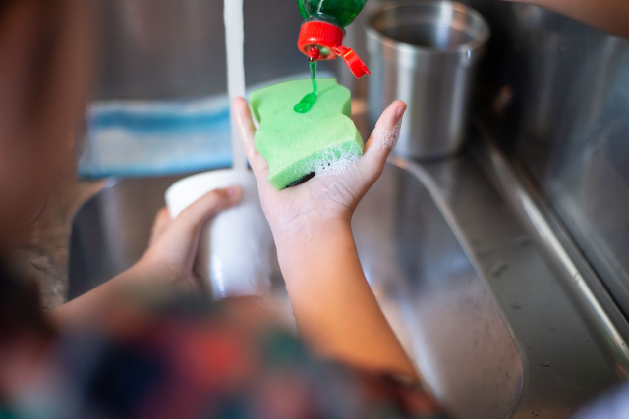 Hands pouring liquid soap onto a sponge under running water in a kitchen sink.